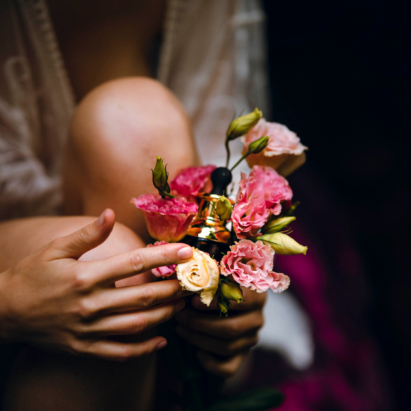 Na zdjęciu zbliżenie na dłonie kobiety, w których trzyma ona bukiet różowych kwiatów. W środek bukietu włożona jest złota buteleczka olejku. Naturalny, ziołowy olejek afrodyzjak Yoniversal Care. Olejek do masażu dla par.
