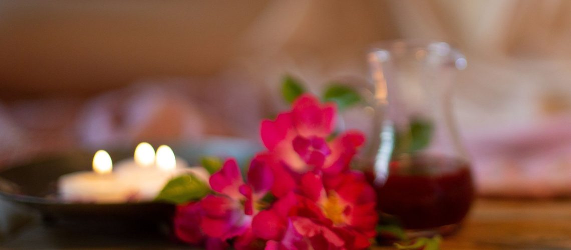 Zdjęcie rozmyte, delikatne, w ciepłym odcieniu. Na zdjęciu bukiecik róż, świeczki oraz dzbanuszek z czerwonym ekstraktem olejowym z dziurawca.
